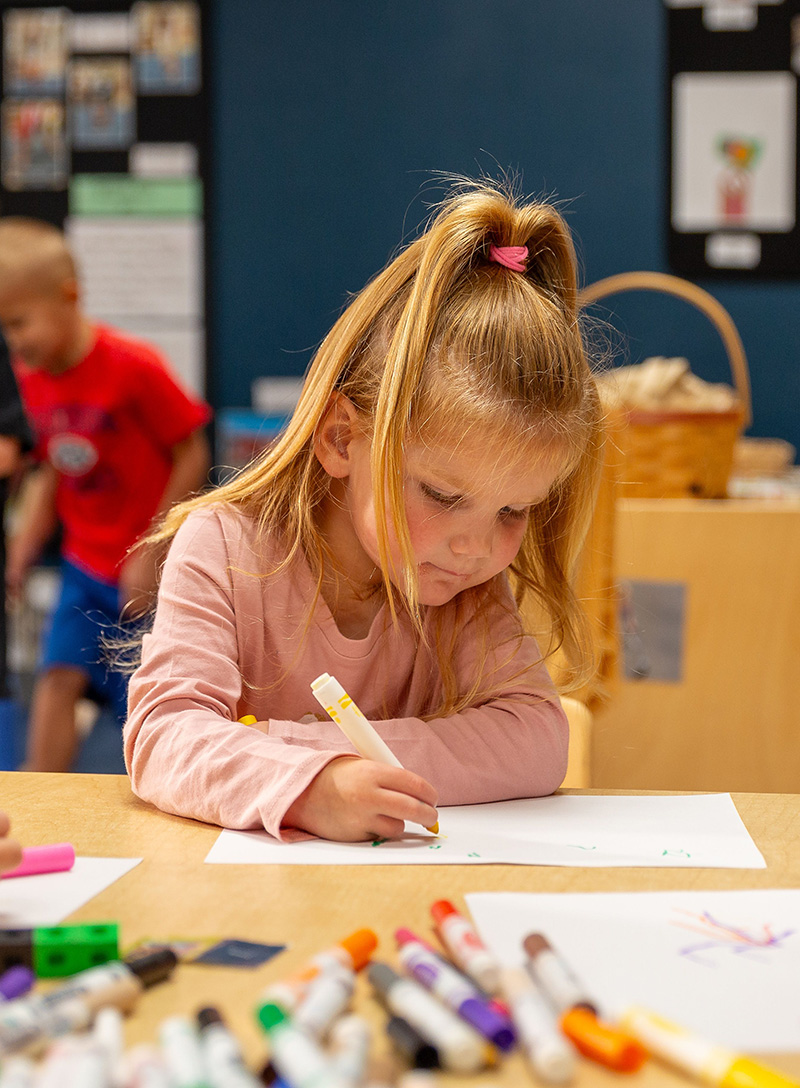 Young girl with long blonde hair in a ponytail sits at a table, focused on drawing with a marker. Colorful markers and art supplies are scattered in front of her.