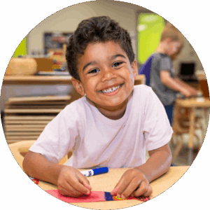 A smiling preschooler sitting at a table and doing a craft with a blue marker.   