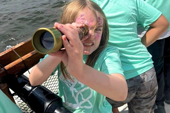 Summer Camp A child wearing a teal summer camp t-shirt and pink face paint looks through a telescope. Camp counselors stand behind them.
