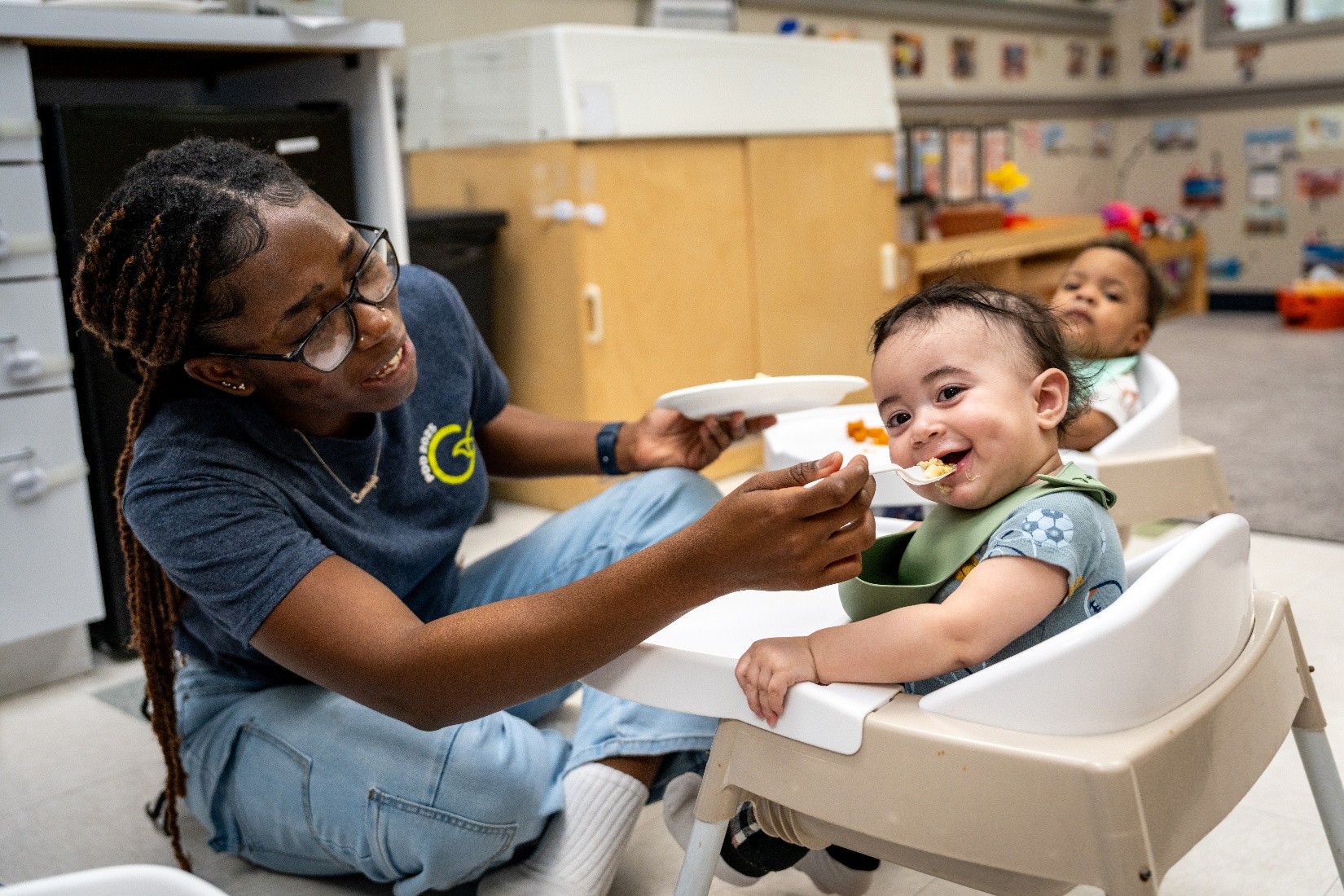 A Celebree School teacher feeding an infant in a high chair who is smiling and wearing a<br />
bib