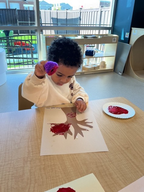Young child creating an apple stamping painting with red paint in a preschool classroom, developing creativity, fine motor skills, and early childhood learning.