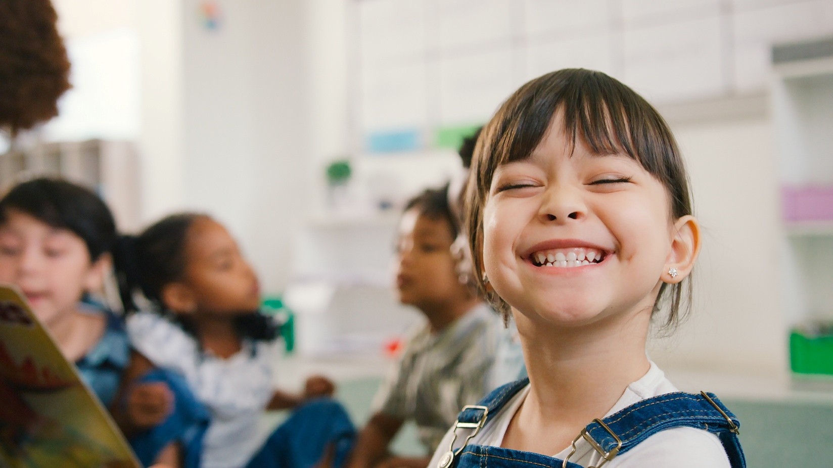 A little girl in pre-K smiling while her classmates learn in the background. 