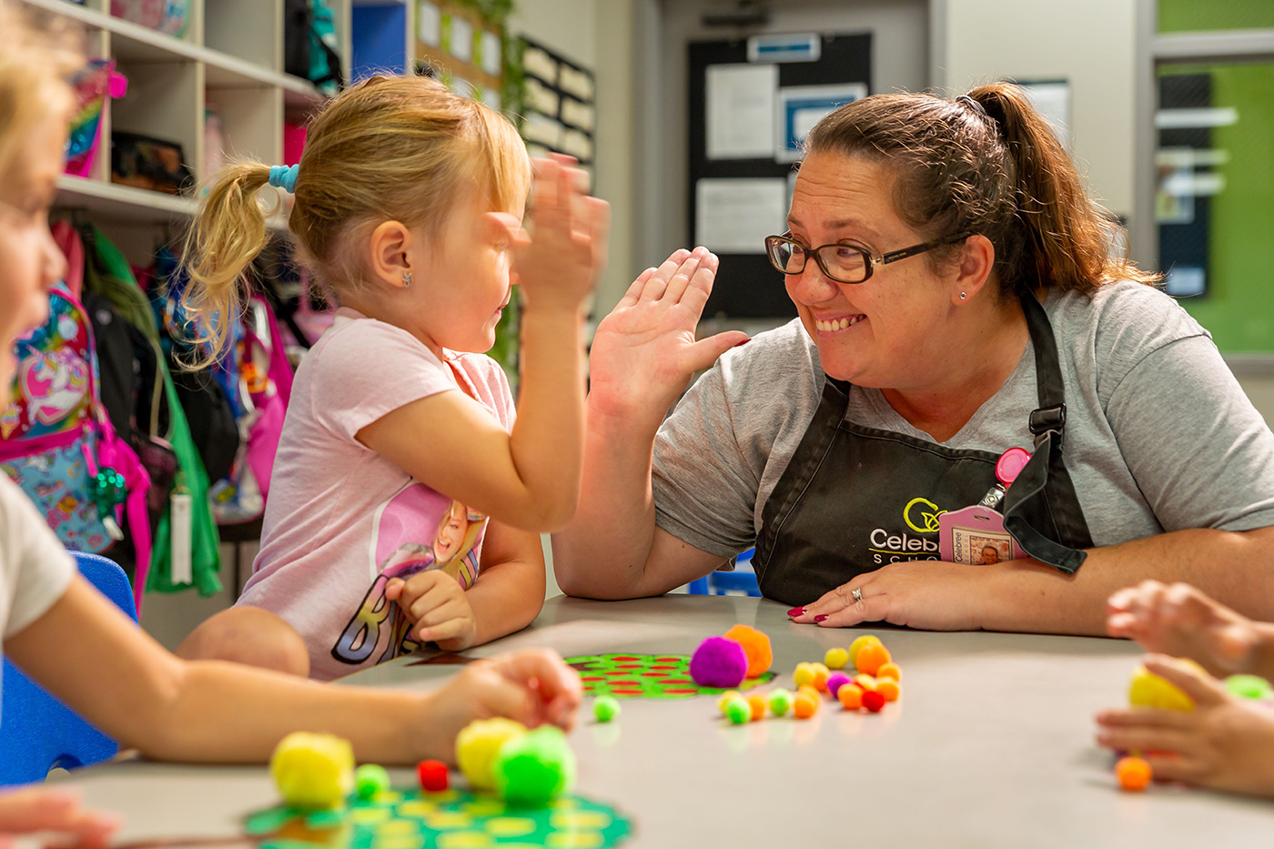 <br />
A female teacher wearing an apron and glasses smiles at a young girl who is raising her hand while they sit at a table with colorful craft materials.