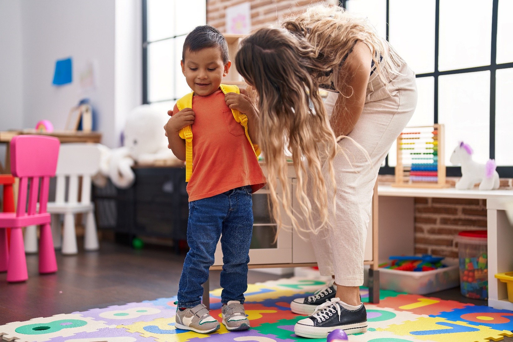 A kid during their first day of day care wearing a yellow back pack while a teacher leans over to say hello. 