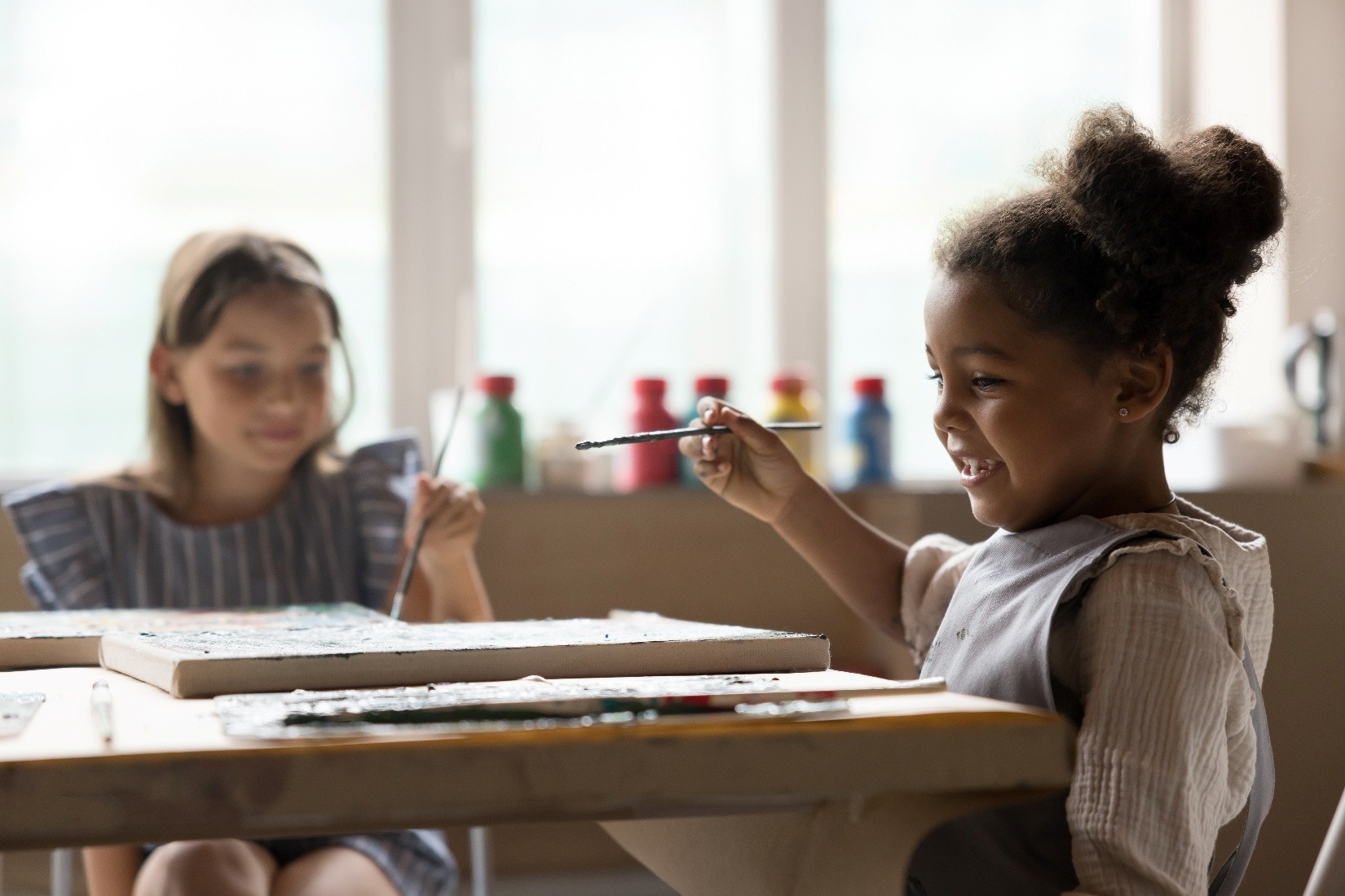 Two little girls in day care sitting at a table and painting while smiling. 