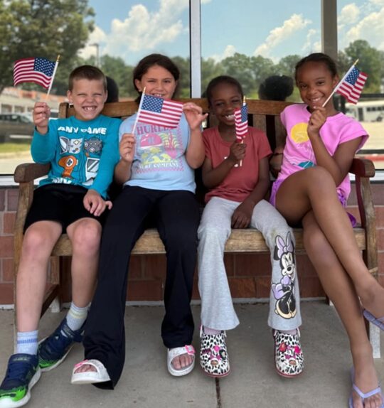 Four smiling children sitting on a bench in front of Celebree School holding American flags during summer camp.