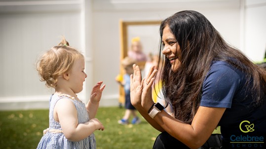 A Celebree School caregiver kneeling down outside in front of a toddler and offering her<br />
hands to give a high five