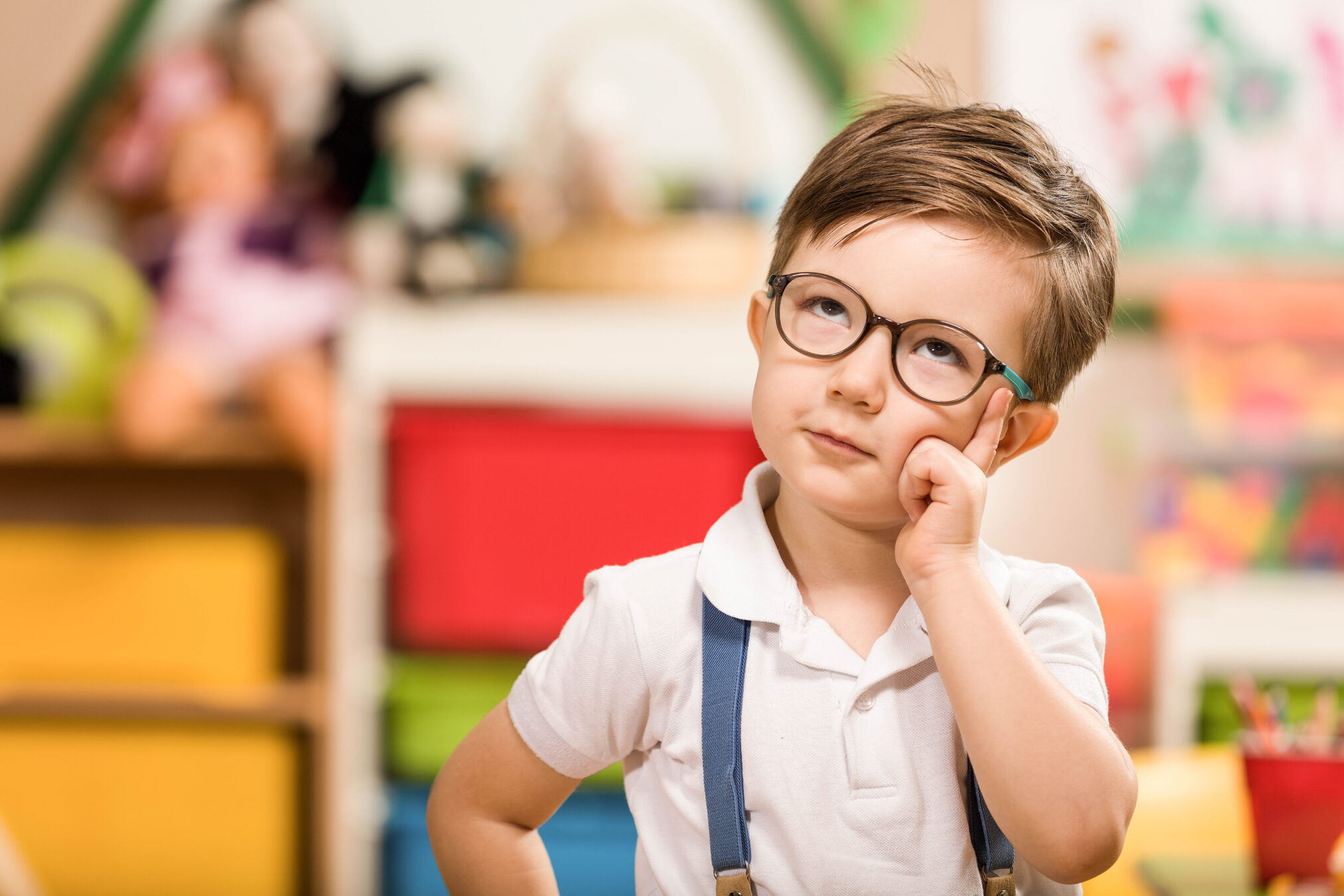 Young preschool boy wearing glasses and suspenders thinking thoughtfully in a colorful classroom, highlighting curiosity and early childhood learning.