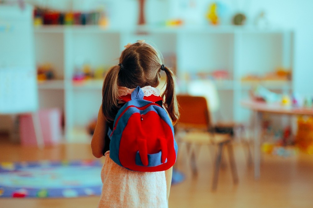 A young girl with a backpack stands in a classroom, looking around with curiosity.
