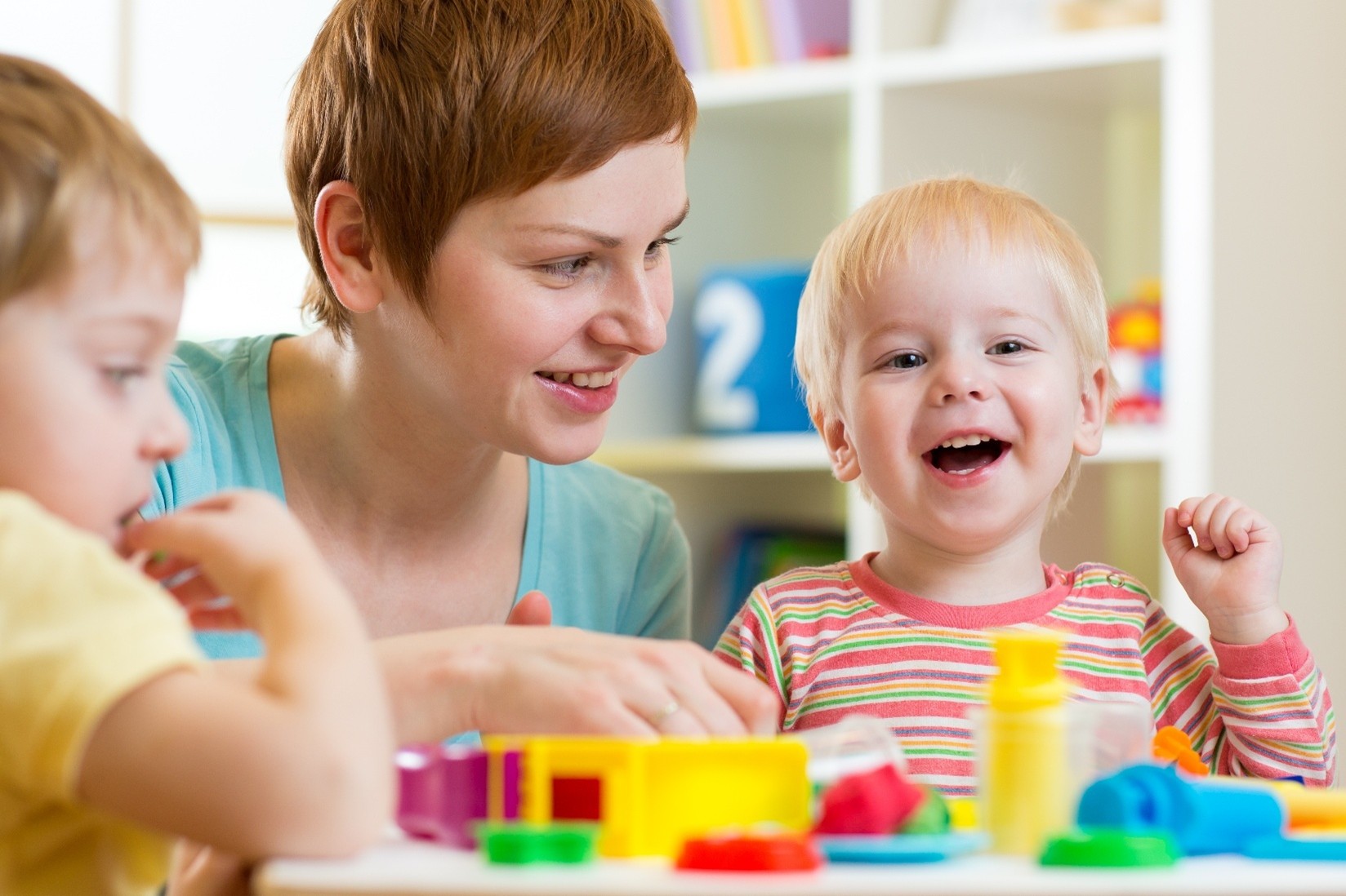 A smiling toddler at day care playing with play dough alongside a teacher and another kid.