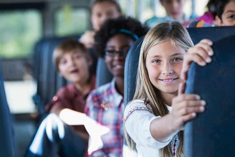 transportation A young girl on a bus smiling with other children smiling behind her.
