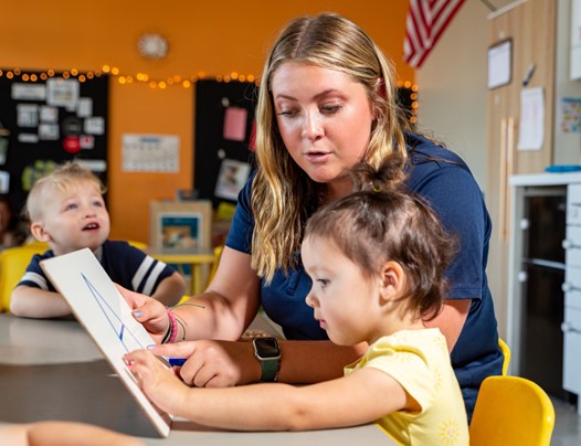 A teacher with blonde hair interacts with two young children at Celebree, pointing at a white board in a colorful classroom.