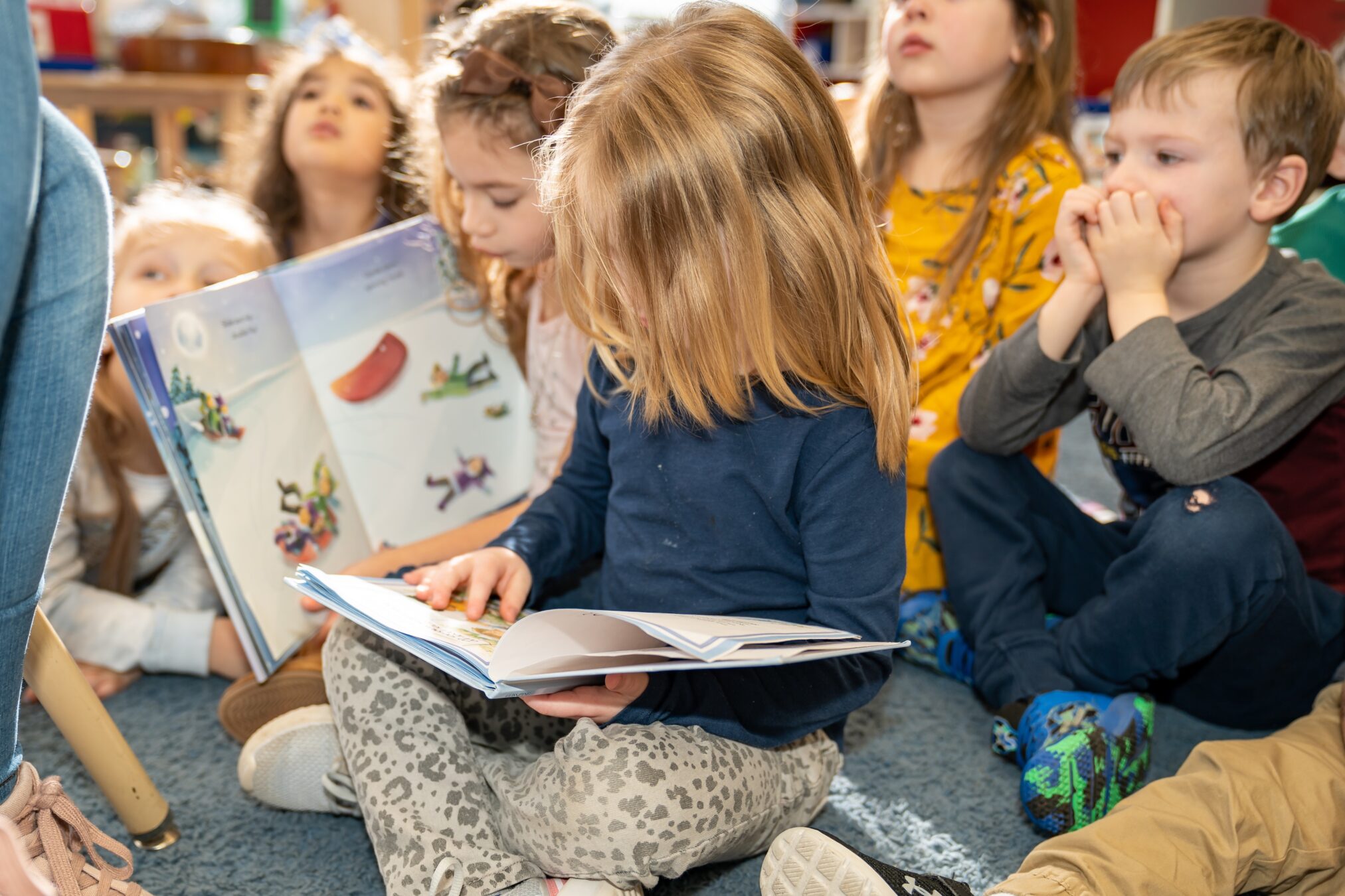 A smiling female preschool teacher sitting on the floor with a group of young children, holding up a number card during an interactive classroom lesson.