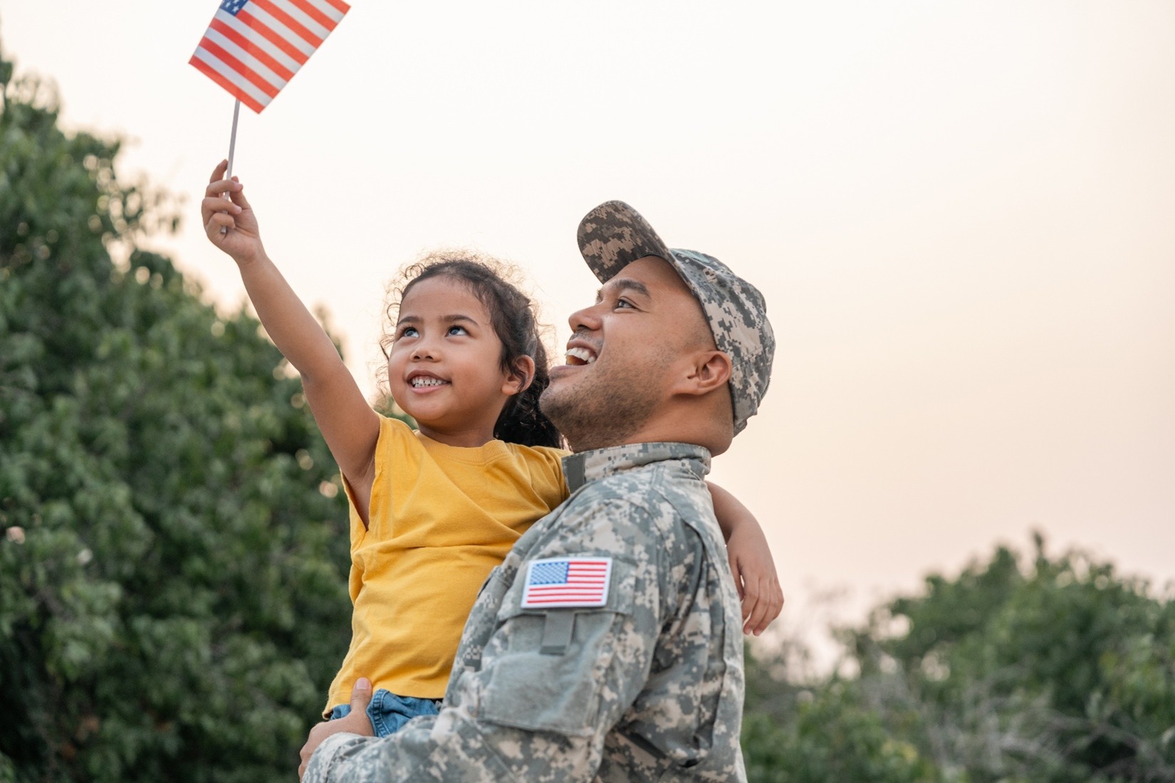 A dad wearing a camouflage military uniform while holding his young daughter as she waves a small American flag in the air.