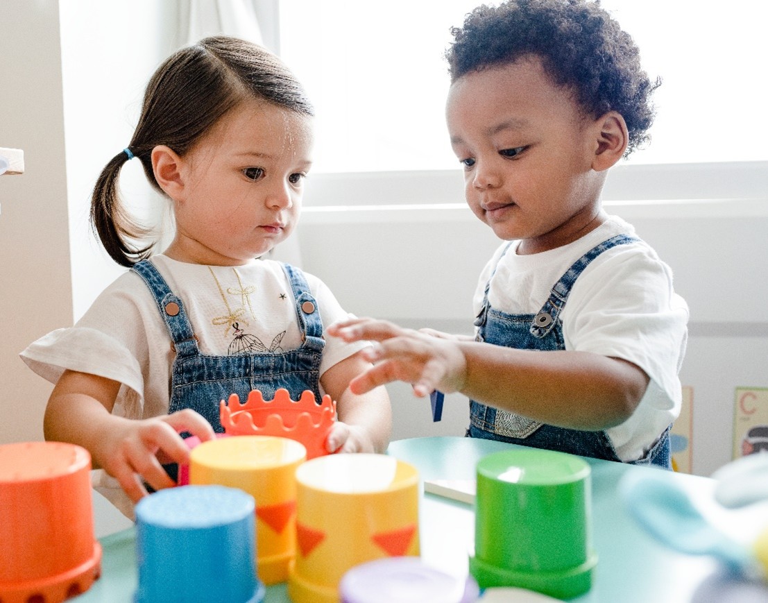 Two young toddlers, one with dark pigtails and the other with curly dark hair, play with colorful stacking cups on a blue table during playtime.
