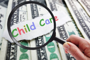 A magnifying glass held over a piece of torn paper that reads "Child Care" in colorful letters, with hundred dollar bills visible underneath.