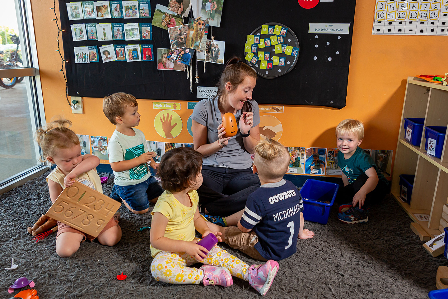 Teacher sitting with four young children on a rug in a classroom, showing them an orange, egg-shaped toy. A blackboard is in the background.