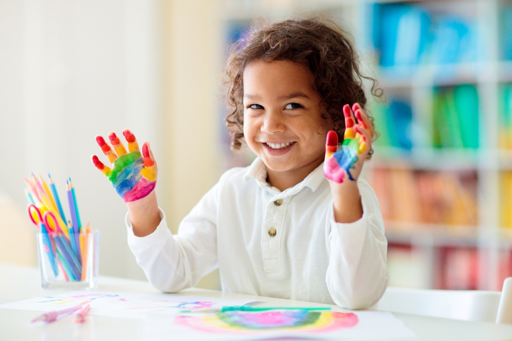 Child Care A child with dark curly hair wearing a white shirt and smiling with rainbow colored paint on their hands.