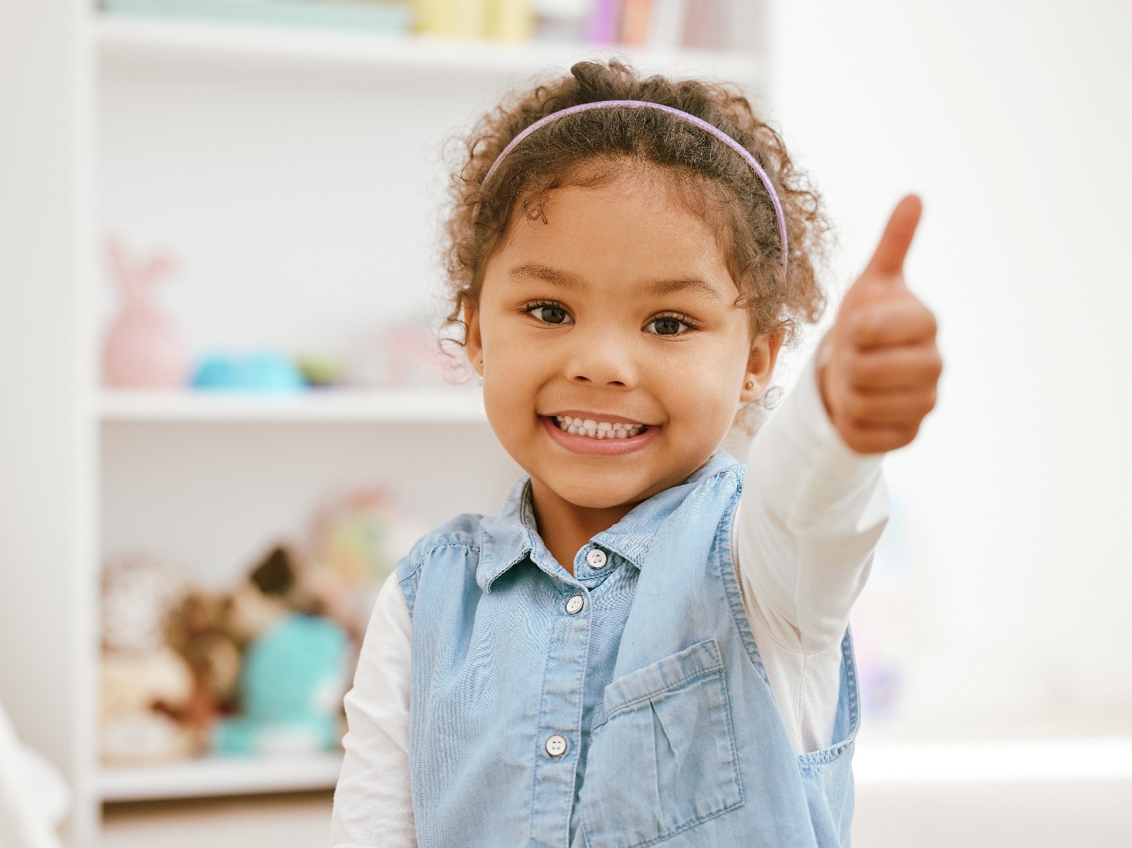 Day Care A little girl with curly hair and a pink headband smiling and giving a thumbs up.