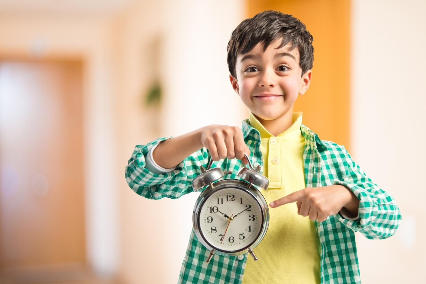 Picture1 (1) A little boy wearing a green gingham shirt over a yellow polo shirt, pointing to a clock and smiling.