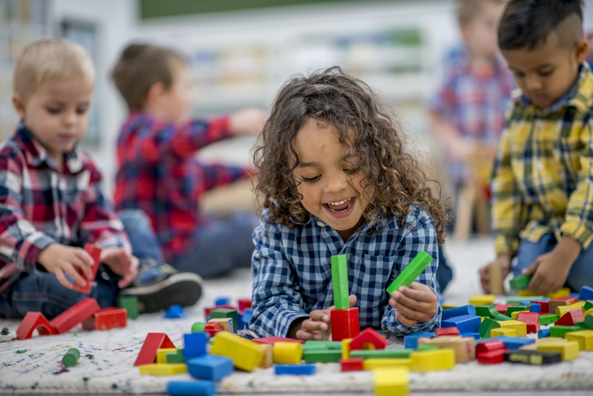 A child with curly hair smiles while playing on the floor with colorful wooden blocks. Other young children are visible in the background also playing with blocks.