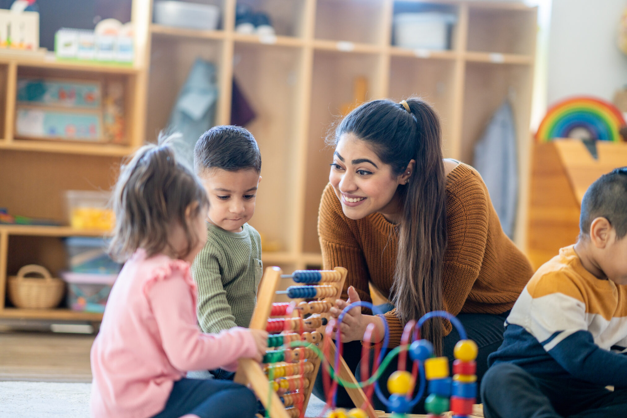 A smiling teacher with long dark hair is kneeling on the floor, helping two young children use a large wooden abacus in a bright classroom.