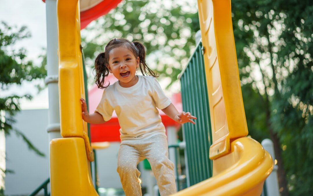 A happy toddler with pigtails about to go down a slide while playing on the playground.