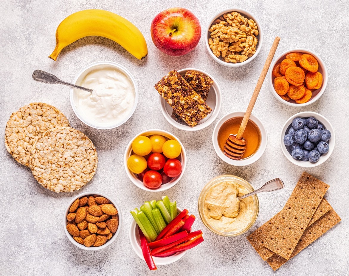 A bird’s eye view of healthy snacks including nuts, vegetables, fruits, honey, hummus, and whole grain snacks. 