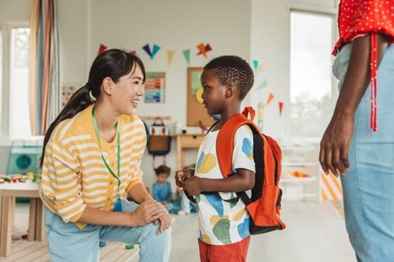 Teacher welcoming a young child with a backpack into a bright daycare classroom while a parent drops them off during their first days at childcare.