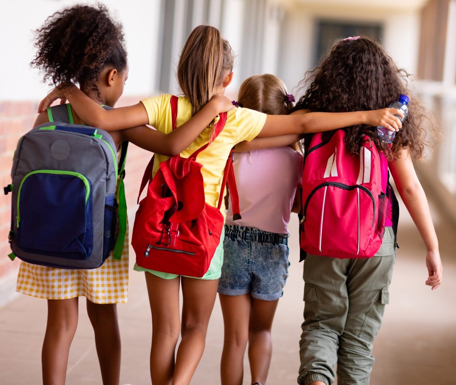 Four girls wearing backpacks and walking with their arms around each other. 