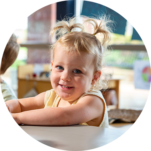 A smiling toddler at a table with blonde hair and pigtails.