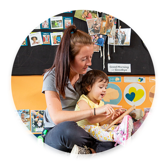 A Celebree School of Grandville caregiver sitting on the floor and reading to a toddler who is sitting in her lap.