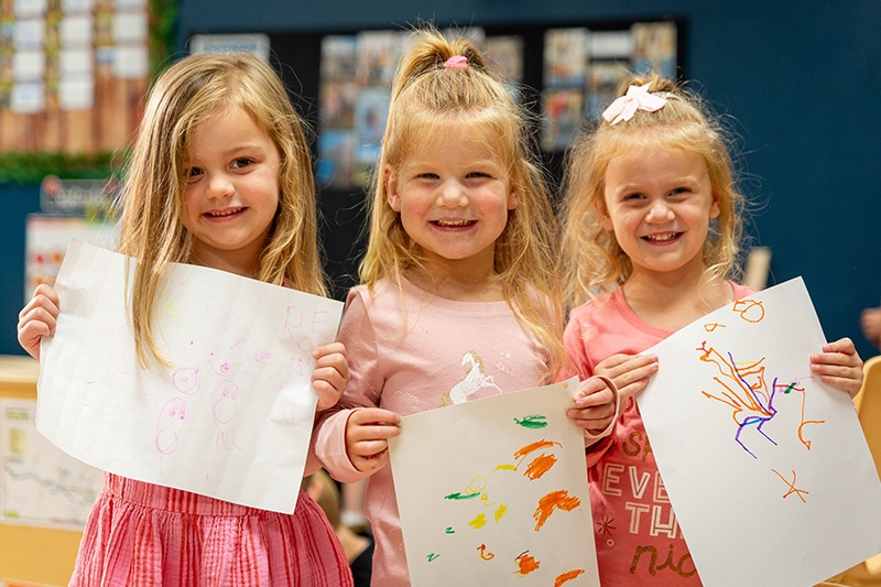 Three little girls with blonde hair smiling and holding their artwork at Celebree School preschool. 