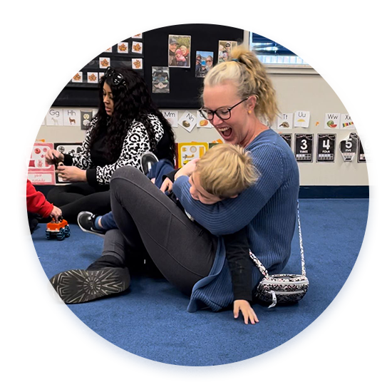 A parent with their child at Celebree School of Grandville holding them and laughing during circle time on the floor.