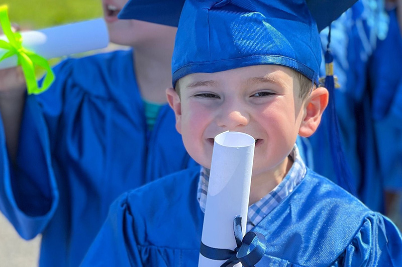 A little boy wearing a blue cap and gown and holding a diploma because he graduated from preschool. 
