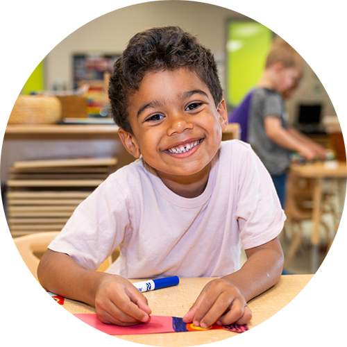 A smiling preschooler sitting at a table and doing a craft with a blue marker.